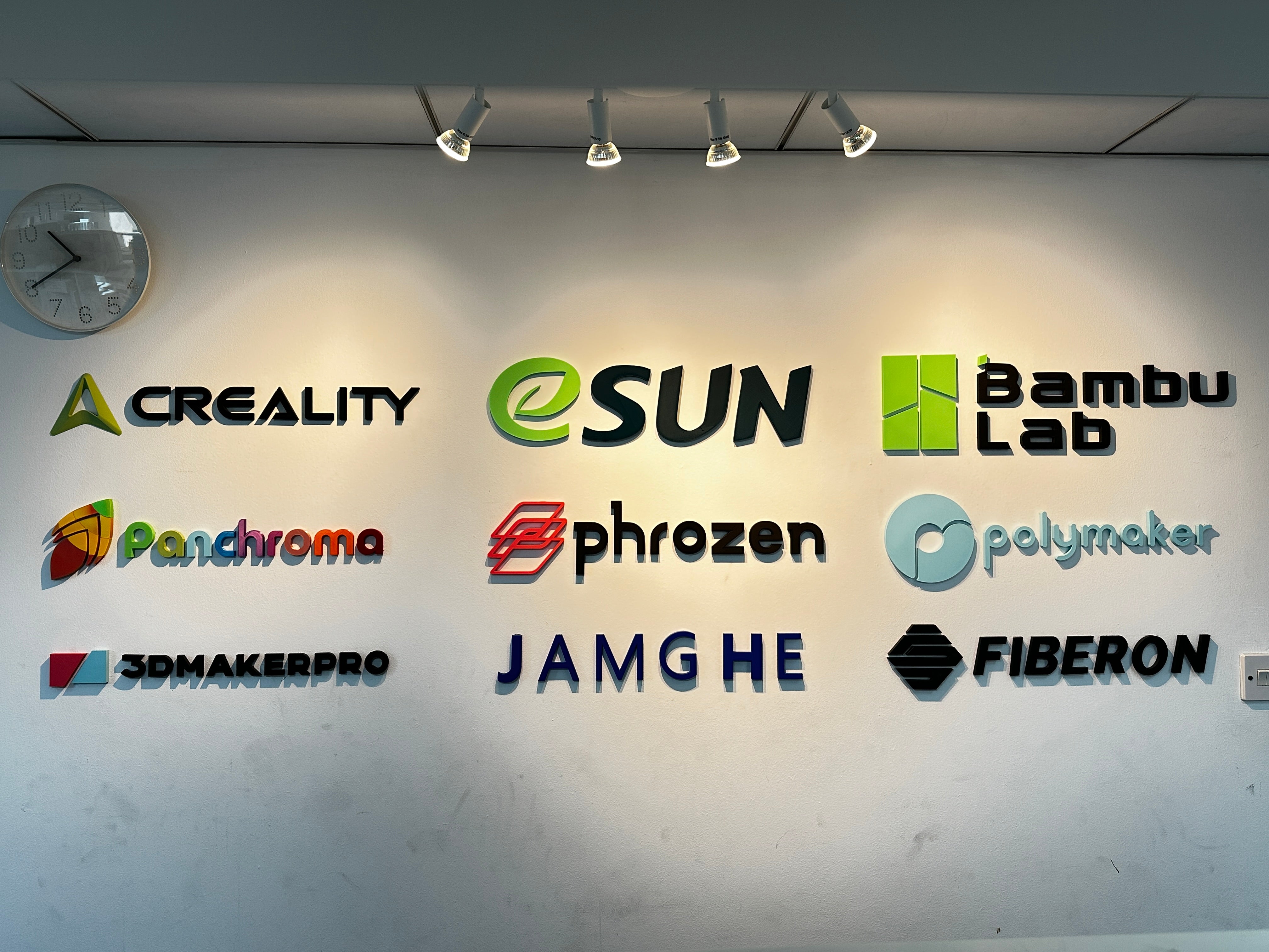Wall display featuring 3D printer and filament brand logos including Bambu Lab, eSUN, Creality, Panchroma, Phrozen, Polymaker, 3DMakerPro, JAMGHE, and Fiberon.